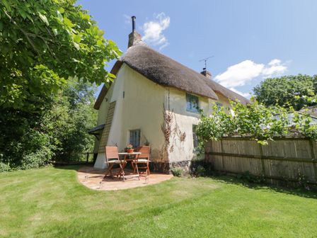A thatched cottage with a small patio table and chairs on a lawn surrounded by trees and a wooden fence at The Hideaway at Burrow Hill in Ottery St Mary