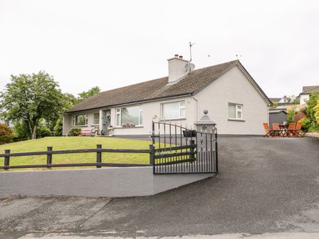 A single story house with a tiled roof a black metal gate a driveway a lawn with a tree and outdoor wooden table and chairs at 27 Pinewood Hill in Warrenpoint Northern Ireland