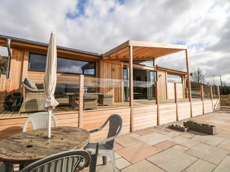 An outdoor patio with a round wooden table and plastic chairs next to a wooden building with glass railing at Stables Lodge in Wingerworth