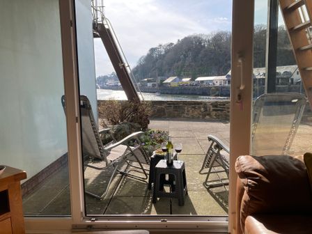 A patio with two reclining chairs and a small table holding a bottle of wine and two glasses overlooking a river with buildings and trees in the background at 48A South Snowdon Wharf in Porthmadog