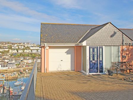 A house with a blue front door garage and a bench overlooking a harbor with boats and houses in the background at Wheal Kendall in Mevagissey