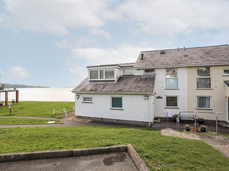 An exterior view of a white and cream two-story building with windows and a sloped roof near a grassy area and waterfront at The Summerhouse in Y Felinheli