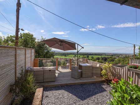 An outdoor patio area with wicker furniture and an umbrella overlooking fields and a village at Aster Cottage in Dyserth
