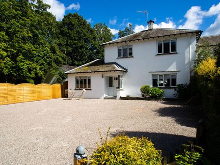 A white two-story house with a gravel driveway and wooden fence surrounded by trees at Bellman Cottage in Bowness-On-Windermere