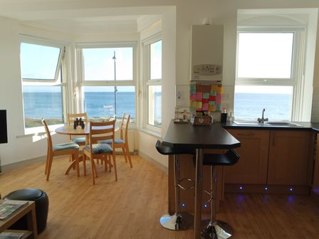 A dining area with a round table and four chairs next to large windows showing a sea view and a kitchen counter with two bar stools at Gwel Y Mor in Pwllheli