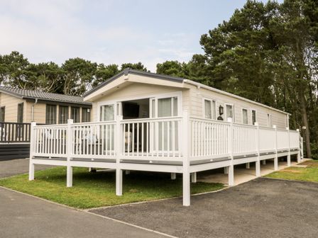A beige mobile home with a white railing deck surrounded by trees at The Rodley Pines in Cayton