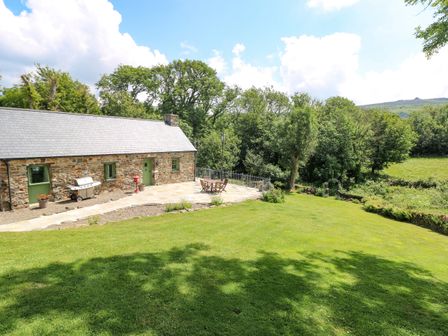 A stone cottage with green doors and windows a barbecue and outdoor furniture on a patio surrounded by grass and trees at Trewrach Cottage in Dinas Cross