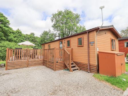 A wooden lodge with a fenced deck and outdoor table with umbrella surrounded by trees at Esthwaite Lodge Beckside 43 Limefitt Park near Windermere