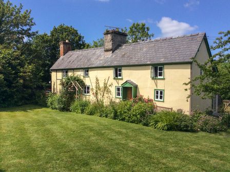 A yellow cottage with green trim surrounded by bushes and a lawn at Denbigh Hall in Presteigne