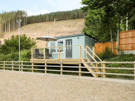 A small cabin with a deck and patio furniture surrounded by trees and a fenced area at Caban Glas in Llanafan near Pontrhydfendigaid