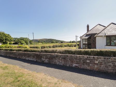 A house with a hedge and brick wall next to a paved path with fields and hills in the background at Laragh in Deganwy