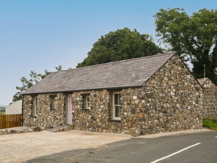A stone cottage with a gray roof and a pink door next to a paved road at Old Shop Cottage in Limavady
