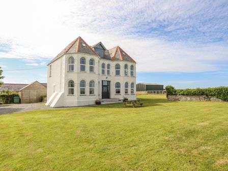 A two-story house with arched windows and a red slate roof on a lawn with a picnic table at Plas Newydd in Llanfaelog near Rhosneigr
