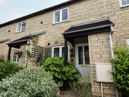 A front view of a stone house with a white door and windows with plants and bushes at Maple Cottage in Broadway