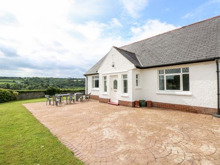 A white house with a tiled patio and plastic chairs and tables surrounded by green lawn at Garth in Pen-y-bryn near Cardigan
