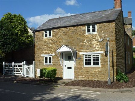 A stone cottage with white windows and door near a white gate and a trimmed bush at Hollytree Cottage in Hook Norton