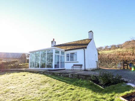 A small white house with a conservatory and a bench outside in a grassy yard at Ty Twmp Tump Cottage in Tal Y Bont On Usk