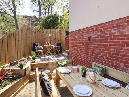 An outdoor patio area with wooden seating and dining table set against a brick and white wall at Coastguards View in Whitby