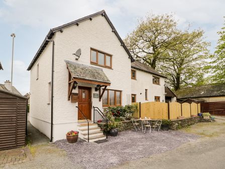 The exterior of a two-story white cottage with wooden door and windows a small outdoor seating area with metal chairs and table and potted plants at Rose Cottage in Burneside