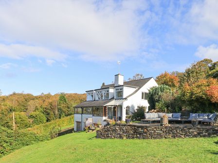 A white two-story house with a patio and outdoor seating area on a grassy hill surrounded by trees at Pheasants Folly in Windermere
