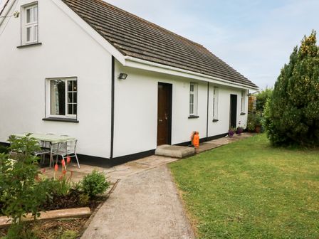 A white house with a tiled roof and a paved walkway leading to a door with a garden area and outdoor table with chairs at Teach Chiuin Coolaheen near Cappoquin County Waterford