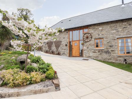 A stone building with wooden windows and door surrounded by a paved driveway and a garden with a blossoming tree at Bagtor Granary in Ilsington