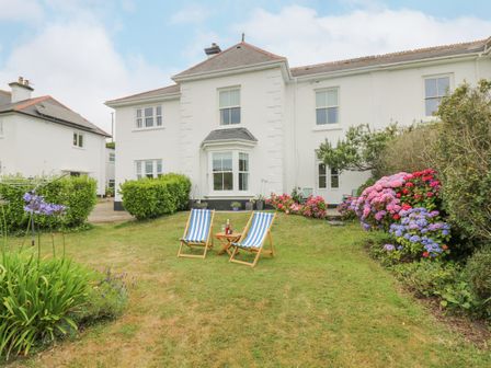 A garden with two striped deck chairs and a small table in front of a white two-story house with flowers and bushes at Chynance in Porthcurno