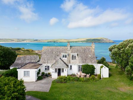 A white house with a stone roof near the sea with green grass and trees at Thalassa in Daymer Bay