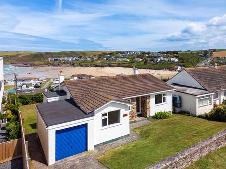 A single-story house with a blue garage door and grassy yard near a beach with houses on hills in the background at Zapadiah in Polzeath
