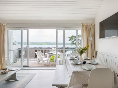 A dining area with a set table and a large window opening to a balcony with white plastic chairs overlooking water at Slipway 10 in Rock