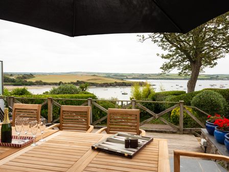 A wooden outdoor table with chairs glasses champagne bottle and backgammon set under a large umbrella overlooking water and fields at Little Strand in Rock