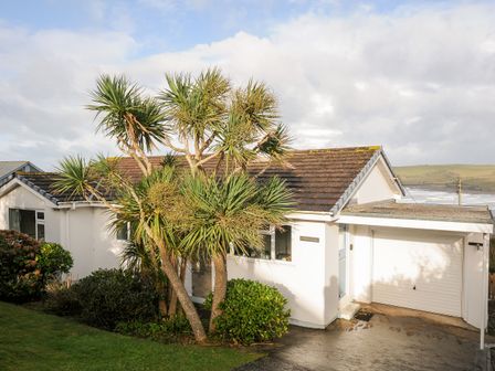 A white house with a tiled roof and attached garage with plants and trees in front at Trevega Polzeath