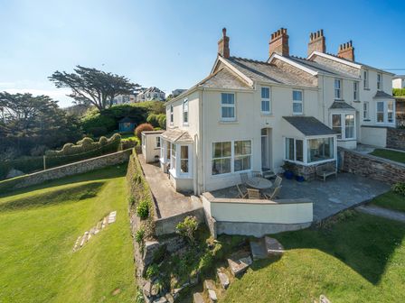 A white two story house with multiple chimneys a stone patio with a round table and chairs and a green lawn at Pentire View 3 Polzeath