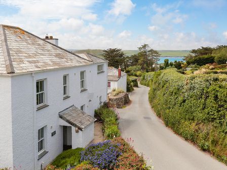 A white house with a slate roof next to a narrow road with green hedges and trees near a body of water at Worthy House in Daymer Bay