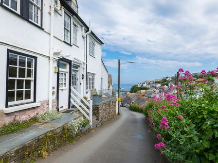 A narrow street with white houses on the left and pink flowers on the right leading to a coastal village and the sea at April Cottage in Port Isaac