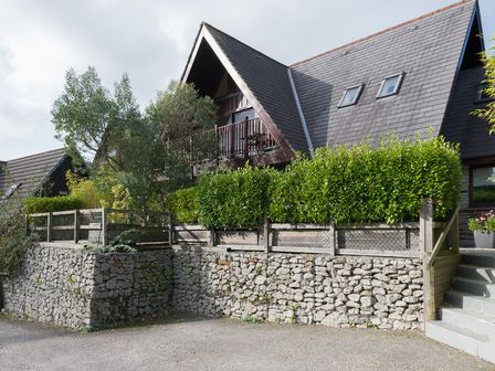 A stone wall with a wooden fence and green shrubs in front of an A-frame house with a balcony at Sandy Hills 8 in Rock