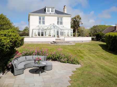 A garden with a patio and curved sofa with table and flowers in front of a large white house at The Haven in Daymer Bay