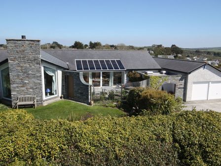 A house with a stone chimney, large window, glass doors, patio furniture, and a hedge in the foreground at Penny Liggan in Rock