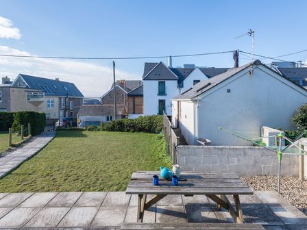 A backyard with grass lawn a wooden picnic table with a teapot and cups on a paved patio and houses in the background at The Holiday House (2 Atlantic Mews) in Polzeath