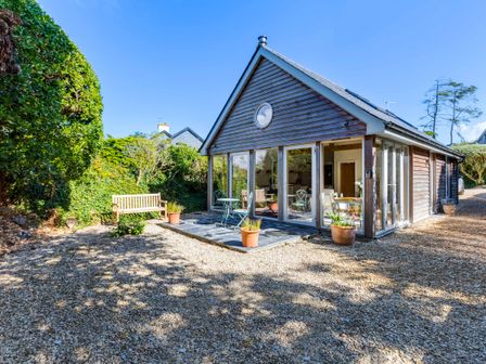 A small wooden house with glass doors and windows a stone patio with metal chairs and table wooden bench and potted plants on a gravel yard at Greystones Studio in Daymer Bay