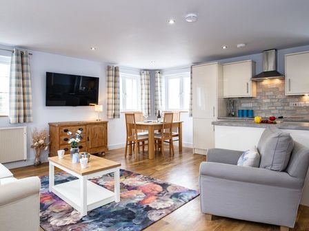 An open plan living and dining area with a wooden dining table and chairs a grey armchair a floral rug a wooden cabinet and kitchen units at The Hideaway in Port Isaac