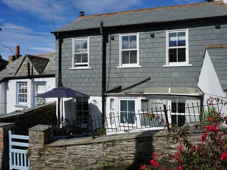 Exterior of a two storey house with gray siding white windows a small patio with table and umbrella and a stone wall in front at Kipper Cottage Port Isaac