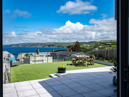 A patio with tiled flooring leading to a grassy area with outdoor wooden furniture and ocean view at Seahaven in Port Isaac