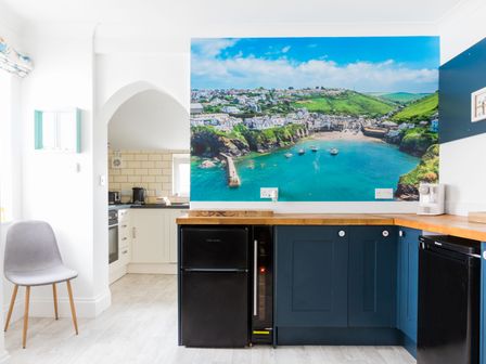 A kitchen with blue cabinets wooden countertops a black fridge a grey chair and a wall mural of a coastal town at Number 28 in Port Isaac
