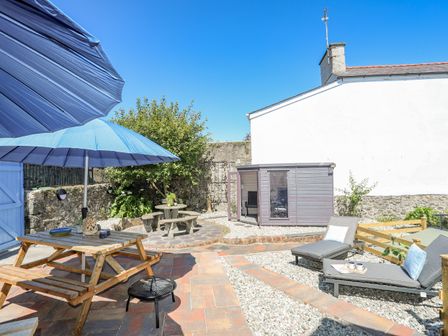 An outdoor patio area with a wooden picnic table under blue umbrellas two lounge chairs with cushions a small round stone table with benches a gray garden shed and stone walls at Gwynle Criccieth
