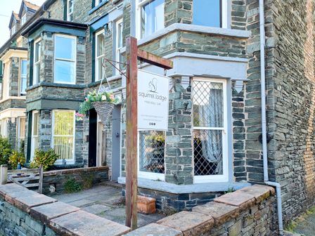 The exterior of a stone building with windows and a hanging basket of flowers a wooden sign for squirrel lodge holiday let at Squirrel Lodge in Keswick