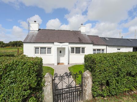 A white cottage with two chimneys and a slate roof behind a black wrought iron gate and hedges at Garnedd Rhos in Brynteg near Benllech