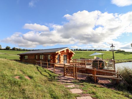 A log cabin with a wooden deck and outdoor furniture next to a lake in a grassy field near Clifton Upon Teme