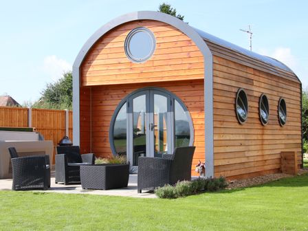 A wooden curved cabin with round windows and black outdoor chairs on a patio with green grass at Cherry Blossom in Diddlebury near Ludlow