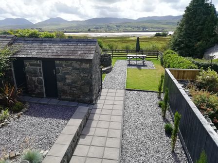 A garden with a stone shed a paved walkway gravel areas small trees a picnic table with an umbrella and a view of fields and mountains at Minffordd in Trawsfynydd